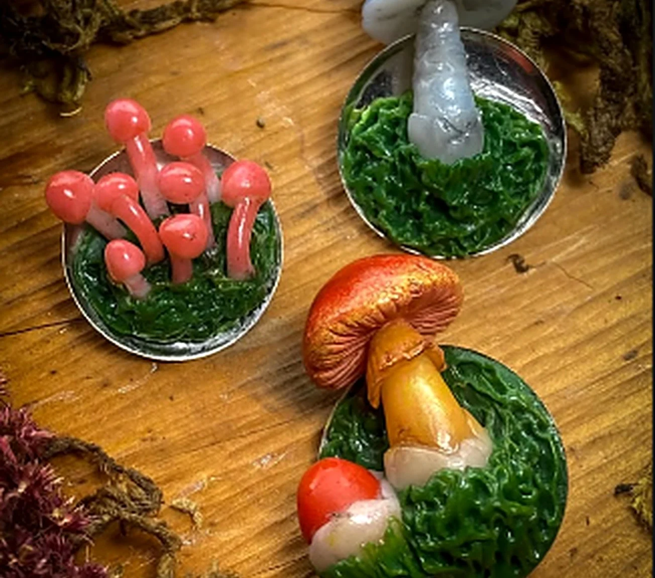 Tres pines de hongos de porcelanicrón sobre una mesa de madera con musgo: un racimo coral, uno gris azulado y uno de sombrero dorado con un hongo rojo pequeño