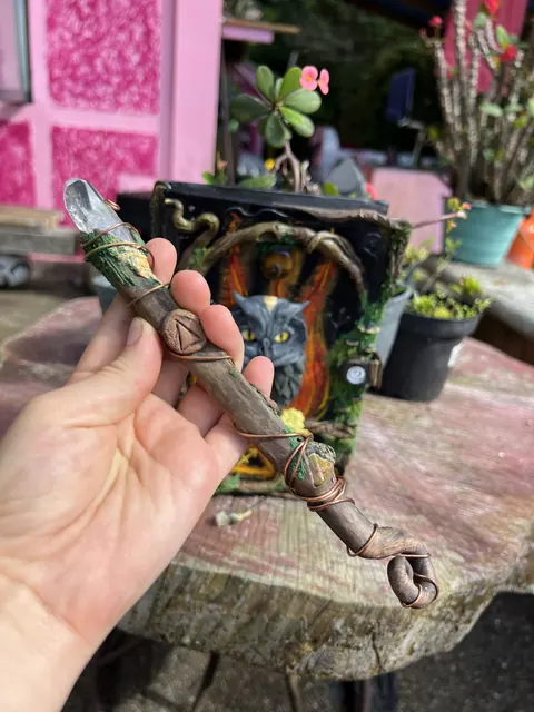 Una mano sostiene una varita de poder con un cristal de cuarzo en la punta, un símbolo de porcelanicrón, musgo y alambre de cobre enroscado